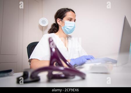 Grave giovane medico donna che indossa uniforme bianca e maschera medica lavorare su un computer portatile con guanti in lattice seduti alla scrivania clinica moderna Foto Stock