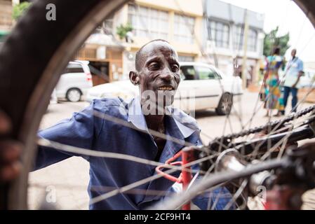 Adulto locale maschio in camicia blu riparazione bicicletta e fissaggio Ruota mentre si siede sulla strada della città in Burundi Africa Foto Stock