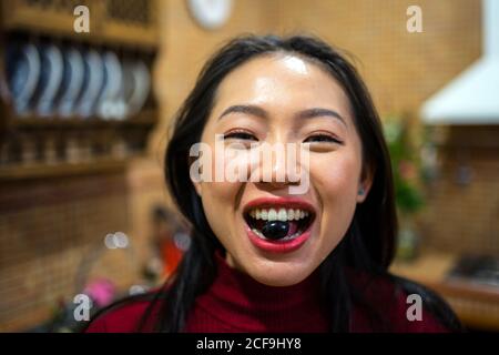 Gioiosa giovane donna asiatica che guarda la macchina fotografica e mostra oliva nera che tiene i denti Foto Stock