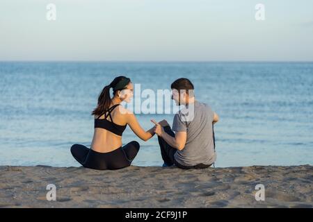 Vista posteriore di giovane coppia allegra multirazziale in abbigliamento sportivo seduto sulla spiaggia di sabbia, riposandosi dopo l'allenamento e godendosi del tempo insieme Foto Stock