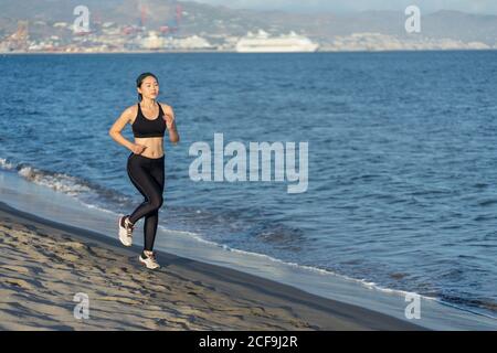 Giovane atleta motivata in abbigliamento nero attivo e sneakers jogging lungo la spiaggia sabbiosa vuota Foto Stock