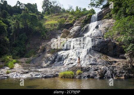 Una forte e schiumosa cascata che scorre da una montagna rocciosa, prima che la piccola Donna si eretti sulle rive del mare in una giornata intensa alle cascate di Diyaluma, Sri Lanka Foto Stock