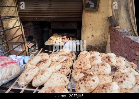 Cairo, Egitto - 14 dicembre 2019: Dall'alto di etnico maschile in cucina casual indossare e vendere tradizionale flatbread seduto al tavolo sulla strada con muro di mattoni che guarda sulla spalla Foto Stock