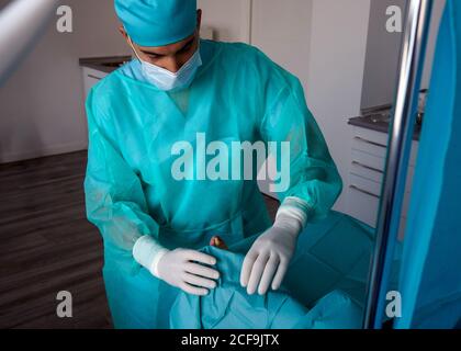 Dall'alto del chirurgo maschile in guanti di gomma uniforme e. maschera sterile preparazione piedi paziente per interventi chirurgici su scavare la punta in ospedale Foto Stock