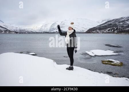 Giovane donna asiatica in giacca calda e cappello che salutano la macchina fotografica mentre si trova in piedi sulla spiaggia contro le bianche e marroni colline innevate e le tranquille acque fresche del lago Kaldfjorden in Norvegia Foto Stock