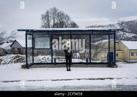 Giovane donna asiatica in abiti caldi guardando il suo orologio in attesa di trasporto a una fermata di autobus da una strada innevata in Norvegia Foto Stock