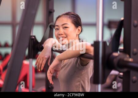 Allegro giovane sportivo etnico in abbigliamento sportivo appoggiato al barbell di metallo della macchina smith e sorridente alla macchina fotografica nel moderno centro fitness Foto Stock