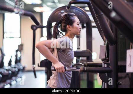 Vista laterale della giovane atleta etnica concentrata in abbigliamento sportivo facendo un'asta si tuffa sulla macchina di esercitazione mentre osservano via dentro moderno centro fitness Foto Stock