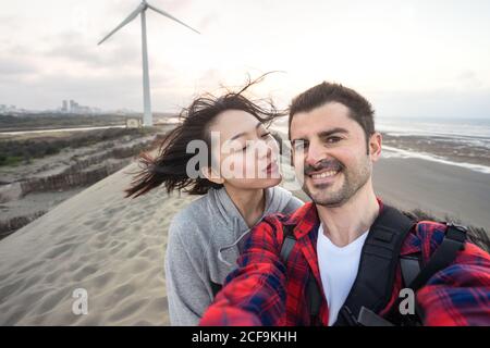 Allegra coppia multirazziale in abbigliamento casual coccolarsi mentre si prende selfie e in piedi sulla sabbia vicino al mare durante il viaggio sotto nuvoloso cielo e guardando la macchina fotografica Foto Stock