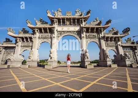 Dal basso di una donna snella che scatta foto con una foto Macchina fotografica mentre si trova sul marciapiede vicino alla facciata del tempio cinese con archi decorati con sculture Foto Stock