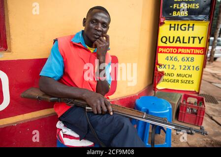 Jinja, Uganda - Settembre, 30:premuroso uomo afroamericano in gilet rosso brillante seduta e custodendo con pistola al negozio con tavola in strada Foto Stock