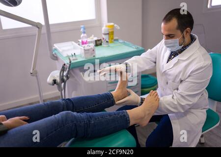 Vista laterale del medico di podiatria maschile in camice bianco e. maschera protettiva massaggiante piedi di paziente femminile in medicina moderna centro Foto Stock