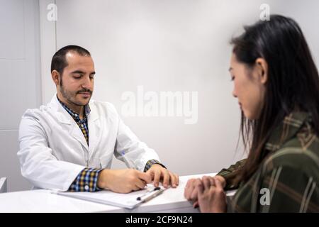 Attento medico maschile che compila il modulo di carta mentre si sta con Paziente femminile asiatica alla reception in ufficio medico Foto Stock