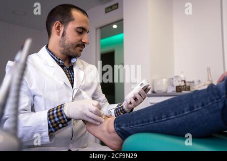 Medico di Podiatria maschile in camice bianco utilizzando attrezzature professionali mentre esame e trattamento dei piedi del paziente in clinica Foto Stock