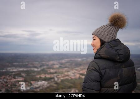 Vista posteriore della donna asiatica sorridente in un cappello a maglia con pompon e giacca calda godendo la città in un posto incredibile a San Marino, Italia Foto Stock