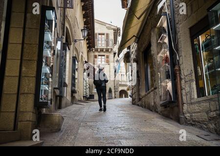 Vista posteriore della donna in giacca calda e cappello camminando lungo la strada guardando attentamente le vetrine dei negozi a San Marino, Italia Foto Stock