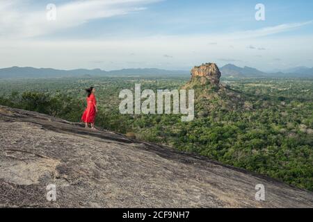 Vista posteriore della donna adulta in vestito rosso godendo di una vista incredibile della fortezza solitaria di roccia Sigiriya nel mezzo dell'altopiano con densa foresta tropicale verde dalla montagna Pidurangala sotto il cielo blu in Sri Lanka Foto Stock