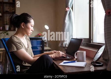 Vista laterale della freelance femminile asiatica in una t-shirt casual e occhiali seduti a tavola e navigando computer mentre si lavora su progetto online a casa Foto Stock