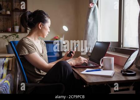 Vista laterale della freelance femminile asiatica in una t-shirt casual seduto al tavolo e navigando il computer che lavora al progetto online a casa mentre si sta inviando messaggi di testo sullo smartphone Foto Stock