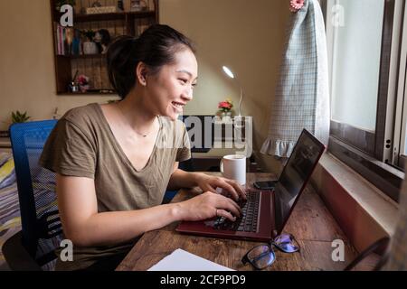 Vista laterale della freelance femminile asiatica in una t-shirt casual e occhiali seduti a tavola e navigando computer mentre si lavora su progetto online a casa Foto Stock