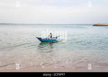 Tangalle, Sri Lanka - 28 luglio 2019: Da sopra vista laterale di giovane pescatore etnico senza volto in abbigliamento casual utilizzando barca tradizionale in mare Foto Stock