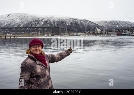 Anziana affascinante donna in vacanza in abiti caldi sorridendo mentre puntando su un mare calmo con ponte e montagne sullo sfondo E guardando la macchina fotografica a Tromso in Norvegia Foto Stock