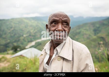 Uganda - 26 novembre 2016: Uomo africano anziano pensivo in abiti casual guardando la macchina fotografica con lo sguardo gentile contro il paesaggio mozzafiato offuscato di verdi colline boschive di giorno Foto Stock