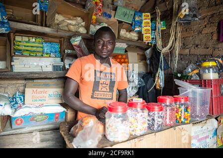 Uganda - Novembre, 26 2016: Uomo africano in t-shirt arancione guardando la macchina fotografica mentre vende vari spuntini nel negozio di strada shabby Foto Stock