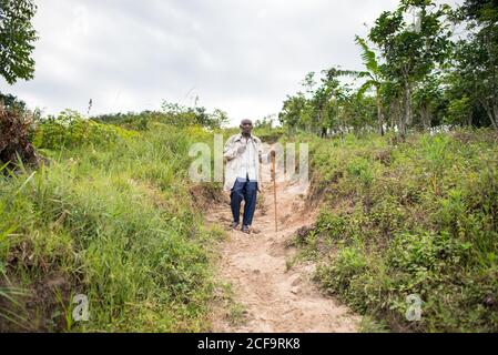Uganda - 26 novembre 2016: Uomo africano anziano pensivo in abiti casual guardando la macchina fotografica con lo sguardo gentile contro il paesaggio mozzafiato offuscato di verdi colline boschive di giorno Foto Stock