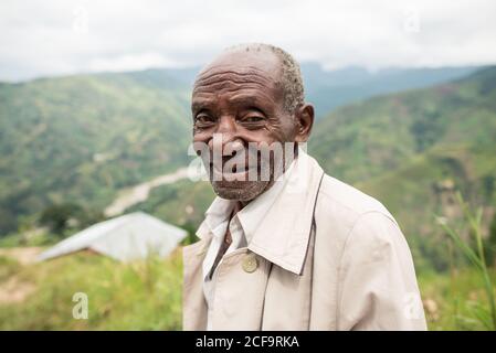Uganda - 26 novembre 2016: Uomo africano anziano pensivo in abiti casual guardando la macchina fotografica con lo sguardo gentile contro il paesaggio mozzafiato offuscato di verdi colline boschive di giorno Foto Stock