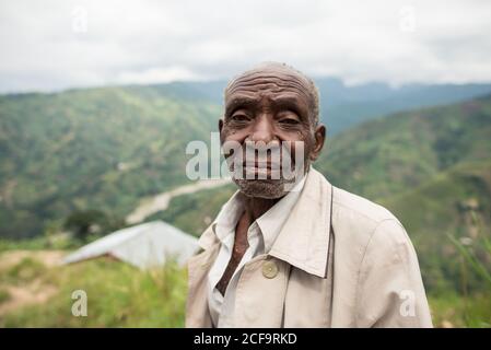 Uganda - 26 novembre 2016: Uomo africano anziano pensivo in abiti casual guardando la macchina fotografica con lo sguardo gentile contro il paesaggio mozzafiato offuscato di verdi colline boschive di giorno Foto Stock