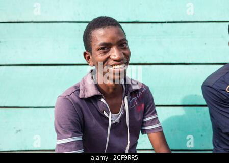 Uganda - 26 novembre 2016: Uomo africano tranquillo in abiti casual guardando la macchina fotografica sorridente seduto al di fuori di una casa di legno blu Foto Stock