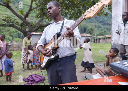 Uganda - 26 novembre 2016: Pensieroso chitarrista africano in camicia bianca guardando via e contemplando mentre si sta accanto al tavolo con attrezzature musicali contro i bambini divertirsi insieme in cortile in campagna Foto Stock