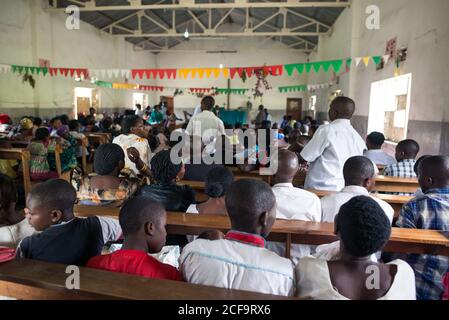 Uganda - 26 novembre 2016: Alto angolo di persone africane adulte e bambini in abiti casual seduti su panchine di legno e ascoltando attentamente il discorso del predicatore durante l'incontro religioso in una spaziosa cappella rustica con pareti in shabby e pavimento in cemento Foto Stock