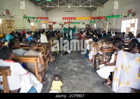 Uganda - 26 novembre 2016: Alto angolo di persone africane adulte e bambini in abiti casual seduti su panchine di legno e ascoltando attentamente il discorso del predicatore durante l'incontro religioso in una spaziosa cappella rustica con pareti in shabby e pavimento in cemento Foto Stock