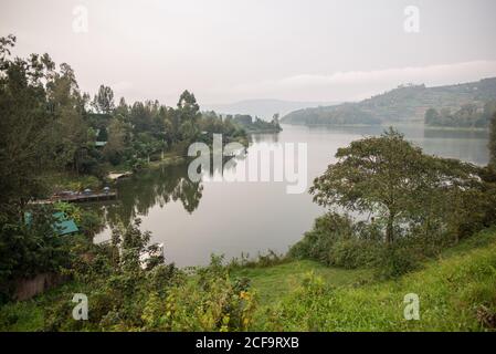 Uganda - 26 novembre 2016: Pittoresca costa selvaggia di un ampio fiume tranquillo con acque tranquille e limpide tra lussureggianti cespugli colorati e verdi alberi decidui contro fossi colline boschive all'orizzonte sotto il cielo nuvoloso di giorno Foto Stock