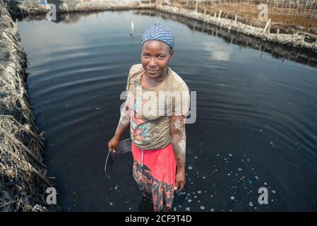 Uganda - 26 novembre 2016: Pensive sorridente donna africana in vividi abiti casual che mette a fuoco e minerando fango nero salato mentre si trova in piedi in acqua scura e utilizzando uno strumento metallico su saline farm contro la vista offuscata di piantagione e cielo blu chiaro in giornata calda Foto Stock