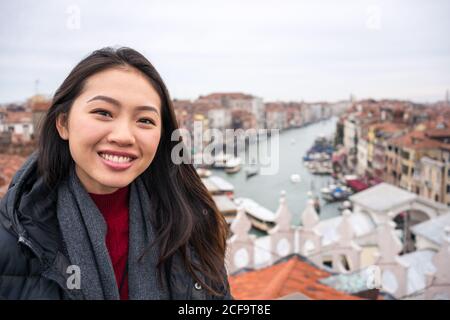 Gioiosa viaggiatore asiatico femminile in abiti caldi sorridente e guardando a macchina fotografica con città antica e corsi d'acqua su sfondo sfocato in tempo nuvoloso Foto Stock