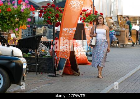 Riga, Lettonia - 21 agosto 2020: Una giovane donna vestita di una parte superiore leggera e una gonna colorata cammina lungo la strada pedonale lungo i caffè di strada e. Foto Stock