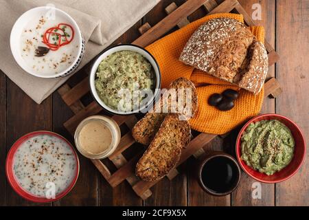 Dall'alto appetitando diverse salse in ciotole rosse bianche con condimenti di semi di sesamo e pane tostato appena sfornato, fragrante e a fette su superficie di legno Foto Stock