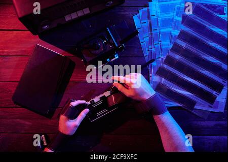 Dall'alto del fotografo irriconoscibile con le mani avvolte in film seduto al tavolo con fotocamere vintage in monolocale scuro Foto Stock