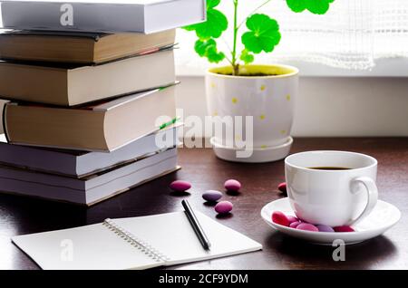 Un concetto accogliente. Una tazza di caffè con cioccolatini multicolore si trova su un tavolo di legno accanto a una pila di libri antichi Foto Stock