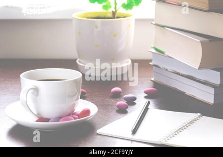 Un concetto accogliente. Una tazza di caffè con cioccolatini multicolore si trova su un tavolo di legno accanto a una pila di libri antichi Foto Stock