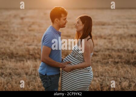 Vista laterale dell'uomo che tocca delicatamente il ventre di donna incinta mentre si guarda l'un l'altro in campo asciutto durante il tramonto in campagna Foto Stock