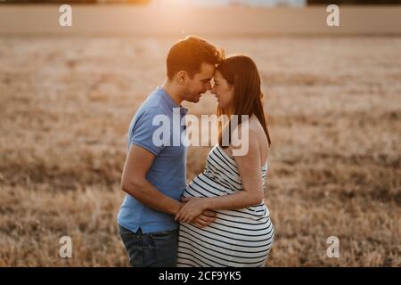 Vista laterale dell'uomo che tocca delicatamente il ventre di donna incinta mentre si guarda l'un l'altro in campo asciutto durante il tramonto in campagna Foto Stock