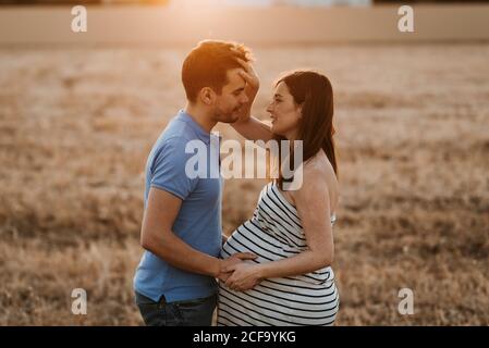 Vista laterale dell'uomo che tocca delicatamente il ventre di donna incinta mentre si guarda l'un l'altro in campo asciutto durante il tramonto in campagna Foto Stock
