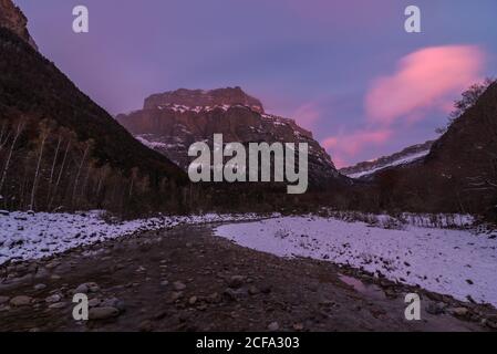 Piccolo ruscello stretto nel mezzo di alberi sottili che corrono attraverso valle di montagna coperta parzialmente di neve sotto pittoresca nuvolosa cielo rosa e viola alla notte in inverno Foto Stock