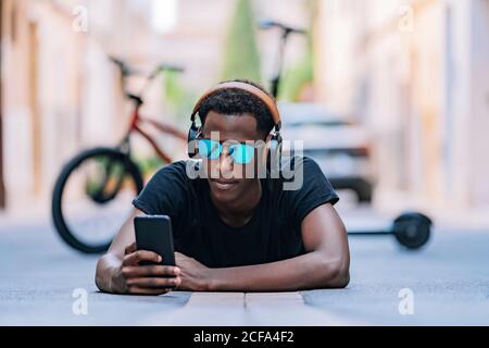 Concentrato giovane afroamericano uomo in occhiali da sole con cuffie e. ascolto di musica sul telefono cellulare mentre si è sdraiati strada asfaltata in strada Foto Stock