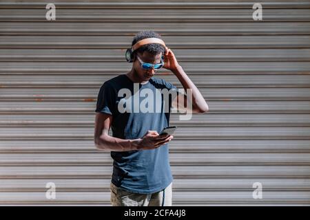 Uomo moderno ed elegante afro-americano in occhiali da sole e cuffie ascolto alla musica sul telefono cellulare alla parete a strisce via Foto Stock