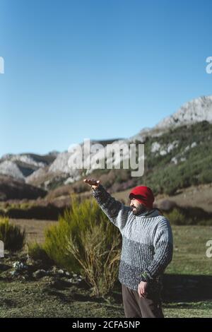 Vista laterale del viaggiatore maschile adulto in pullover grigio e cappellino rosso lavorato a maglia in piedi con la mano rialzata sulla montagna valle e guardando in lontananza in giornata di sole con blu cielo Foto Stock